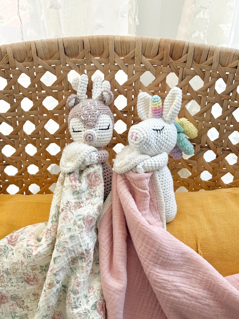 May include: Two crochet security blankets featuring a deer and a unicorn. The deer has a beige head with antlers and a floral print blanket. The unicorn has a white head with a rainbow horn and mane, paired with a solid light pink muslin blanket.