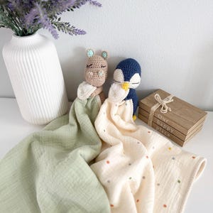May include: Two crochet security blankets featuring a tan hippo and a navy blue penguin head. The hippo is attached to a solid sage green muslin cloth, while the penguin is attached to a cream muslin cloth with small multicolored polka dots.