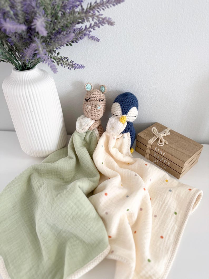May include: Two crochet security blankets featuring a tan hippo and a navy blue penguin head. The hippo is attached to a solid sage green muslin cloth, while the penguin is attached to a cream muslin cloth with small multicolored polka dots.