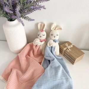 May include: Two crochet rabbit security blankets with cream-colored heads and embroidered closed eyes. One rabbit has a pink flower and a pink muslin blanket, while the other has a blue neck accent and a blue muslin blanket with cream trim.