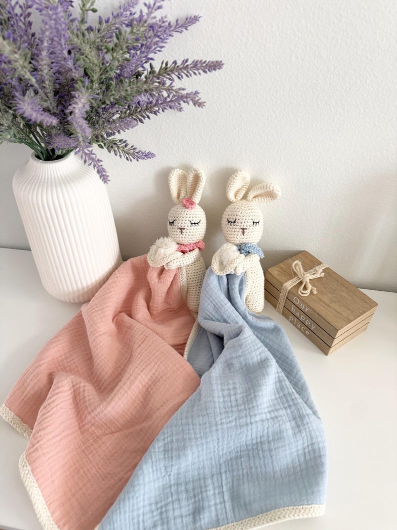 May include: Two crochet rabbit security blankets with cream-colored heads and embroidered closed eyes. One rabbit has a pink flower and a pink muslin blanket, while the other has a blue neck accent and a blue muslin blanket with cream trim.
