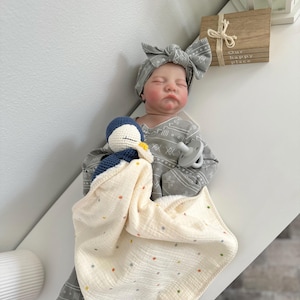 May include: A realistic baby doll with closed eyes, wearing a gray long-sleeved outfit with a white geometric pattern and a matching knotted headband. The doll holds a small blue and white crocheted penguin toy attached to a cream-colored muslin blanket.