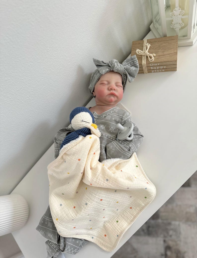 May include: A realistic baby doll with closed eyes, wearing a gray long-sleeved outfit with a white geometric pattern and a matching knotted headband. The doll holds a small blue and white crocheted penguin toy attached to a cream-colored muslin blanket.