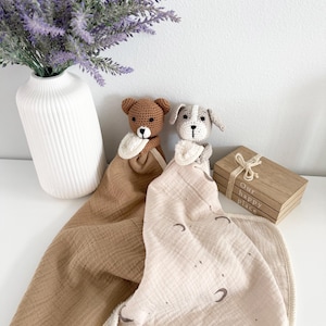 May include: Two crochet security blankets featuring a brown bear head and a grey dog head. The bear blanket is solid brown muslin, while the dog blanket is cream muslin with a brown moon and star print. Both have cream-colored trim and small bibs.