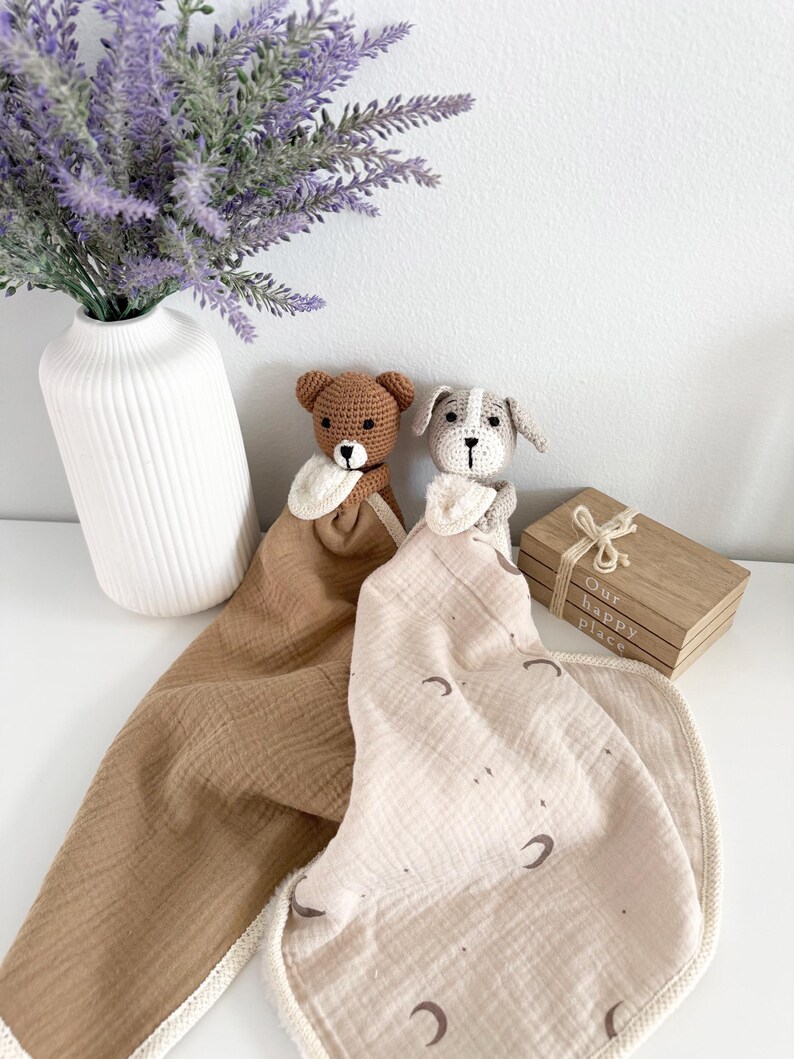May include: Two crochet security blankets featuring a brown bear head and a grey dog head. The bear blanket is solid brown muslin, while the dog blanket is cream muslin with a brown moon and star print. Both have cream-colored trim and small bibs.
