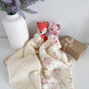 May include: Two crochet security blankets featuring a stuffed fox head and a stuffed pig head. The fox is attached to a solid cream-colored muslin cloth, while the pig is attached to a cream-colored muslin cloth with a pink and green floral pattern.