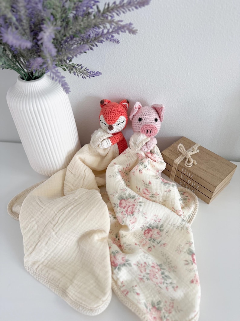 May include: Two crochet security blankets featuring a stuffed fox head and a stuffed pig head. The fox is attached to a solid cream-colored muslin cloth, while the pig is attached to a cream-colored muslin cloth with a pink and green floral pattern.
