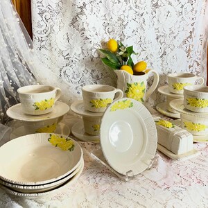 May include: A vintage cream-colored ceramic tea set with yellow floral accents. The set includes cups, saucers, bowls, a creamer, and a butter dish. The floral design is repeated on each piece. The set is arranged on a white lace tablecloth.