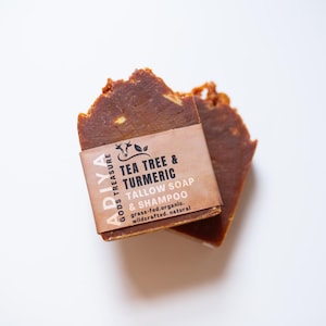 May include: Two bars of brown soap with a paper label that reads "Tea Tree & Turmeric Tallow Soap & Shampoo." The soap is a reddish-brown color and has a rough, uneven texture. The label also includes the brand name "Adiya Gods Treasure."