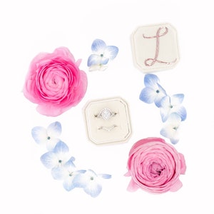 May include: A white ring box with two rings inside, surrounded by pink and blue flowers. The box has a gold letter "L" on the top.