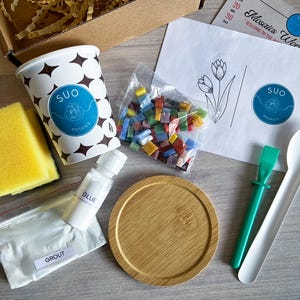 May include: A mosaic tile craft kit with a white paper instruction sheet, a cup of colourful glass tiles, a white cup with the words "SUO MOSAIC" printed on it, a white bowl, a green tool, a white spoon, a white bottle of glue, a white bag of grout, and a yellow sponge.
