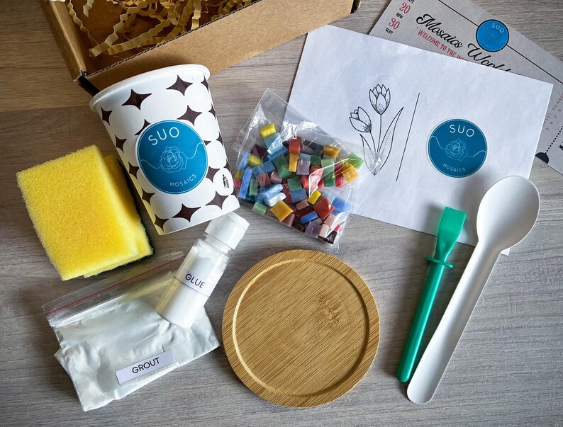 May include: A mosaic tile craft kit with a white paper instruction sheet, a cup of colourful glass tiles, a white cup with the words "SUO MOSAIC" printed on it, a white bowl, a green tool, a white spoon, a white bottle of glue, a white bag of grout, and a yellow sponge.