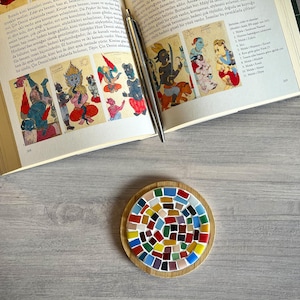 May include: A round wooden coaster featuring a colourful mosaic tile design. The tiles are arranged in a spiral pattern and are in shades of red, blue, green, yellow, and brown.