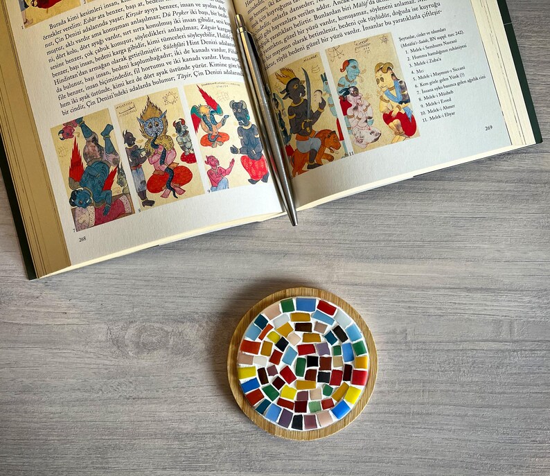 May include: A round wooden coaster featuring a colourful mosaic tile design. The tiles are arranged in a spiral pattern and are in shades of red, blue, green, yellow, and brown.