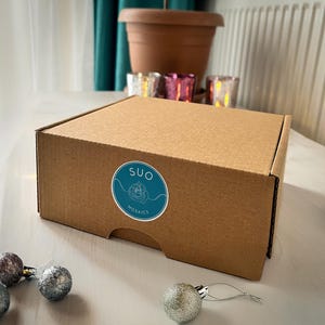 May include: A brown cardboard box with a white sticker that says "SUO Mosaics" in a teal blue circle. The box is sitting on a white surface with silver baubles.