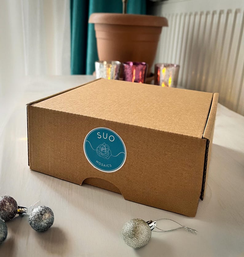 May include: A brown cardboard box with a white sticker that says "SUO Mosaics" in a teal blue circle. The box is sitting on a white surface with silver baubles.
