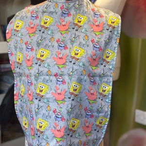 Puede incluir: Un babero gris con un patrón repetido de personajes de dibujos animados, incluyendo Bob Esponja, Patricio Estrella, Calamardo Tentáculos y Don Cangrejo. El babero está hecho de una tela suave y tiene un borde inferior curvo.