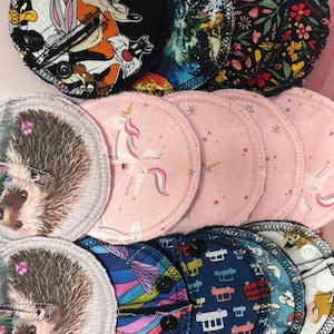 May include: A selection of reusable cloth sanitary pads with diverse, vibrant patterns. Designs include cartoon characters, space scenes, floral prints, hedgehogs, and unicorns. Each pad has a dark trim and a snap closure.