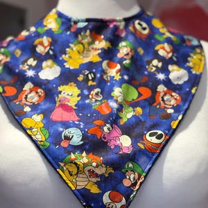 Könnte beinhalten: Ein blauer Lätzchen mit einem farbenfrohen Muster von Super Mario-Charakteren. Das Lätzchen zeigt Mario, Luigi, Prinzessin Peach und Bowser. Es hat einen dunkelblauen Rand und besteht aus einem weichen Stoff, ideal für Babys.