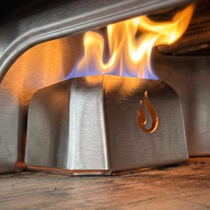 May include: Close-up of a stainless steel pizza oven burner with flames. The burner has a brushed metal finish and a flame-shaped cutout. The flames are orange and yellow, with blue at the base.