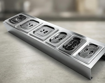 May include: A stainless steel food serving station with six rectangular compartments. Each compartment has a lid with two oval cutouts. The station is angled, with a polished finish, and sits on a light-coloured surface.