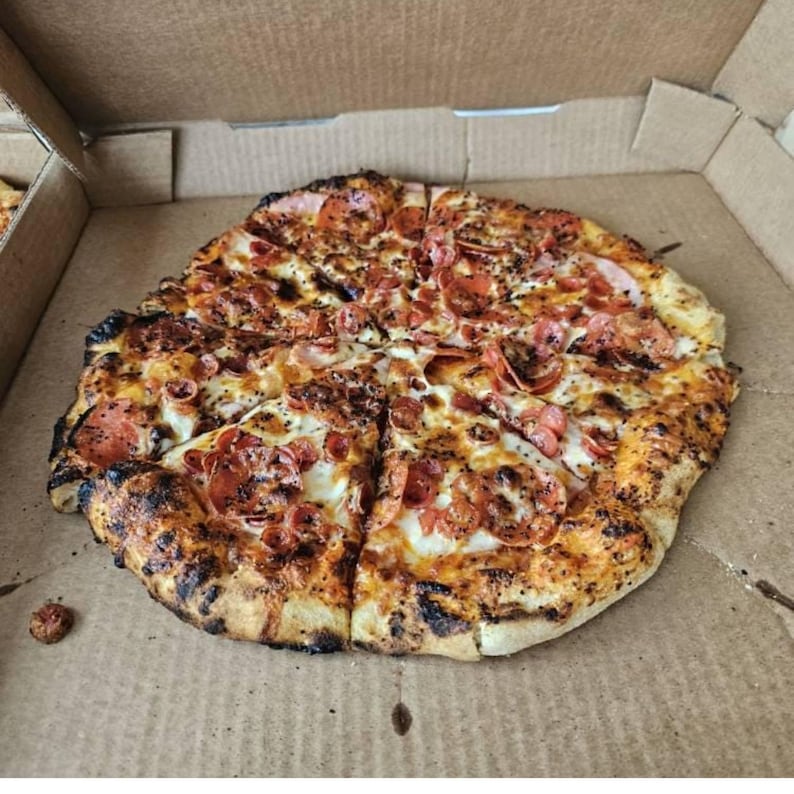 Puede incluir: Una pizza reci&eacute;n horneada con pepperoni y otros ingredientes, cortada en rodajas, dentro de una caja de cart&oacute;n. La corteza es dorada con bordes oscuros y ligeramente carbonizados. Las rodajas de pepperoni est&aacute;n esparcidas por la pizza, con queso derretido y otros ingredientes.