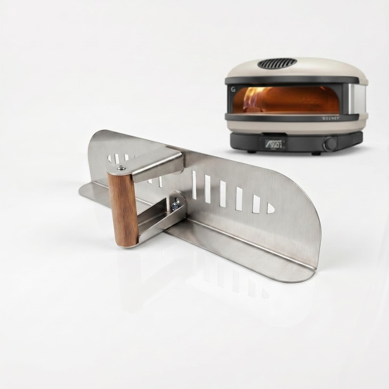 May include: A stainless steel pizza peel with a wooden handle is angled in front of a small, cream-colored pizza oven. The oven has a black base and a digital display. The peel has a series of rectangular cutouts.