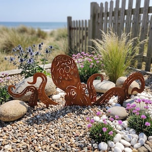 May include: A rust-colored metal octopus sculpture with intricate cut-out circular patterns on its head and textured tentacles, positioned among smooth white stones, green plants, and purple flowers in an outdoor garden setting.