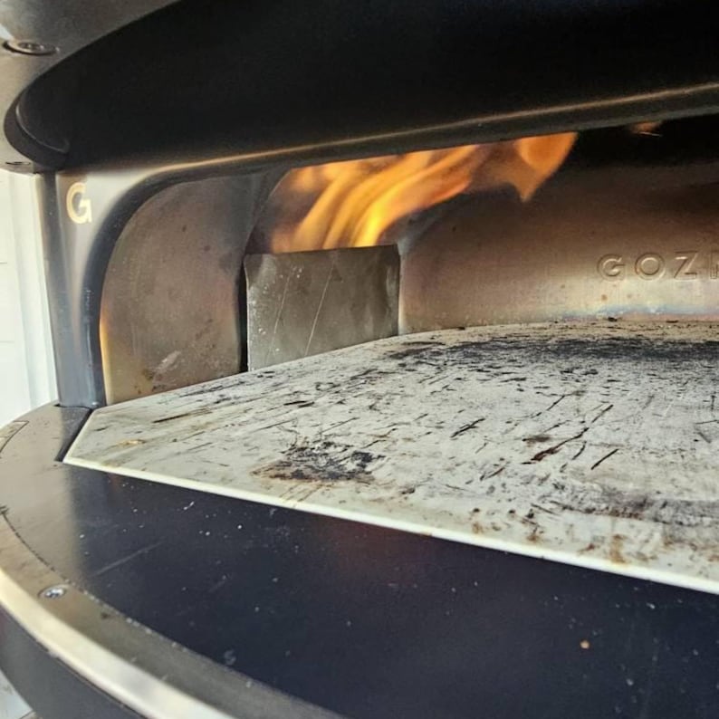Puede incluir: Primer plano del interior de un horno de pizza con llamas visibles. El horno tiene un exterior oscuro y una superficie de piedra de color claro. Las llamas son de color naranja brillante y amarillo. La marca "GOZNEY" es visible en el horno.