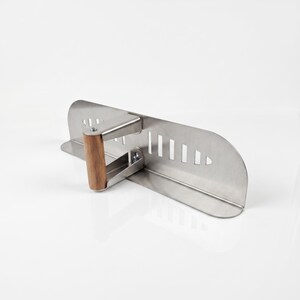 May include: A stainless steel kitchen tool with a wooden handle. The tool has a flat, rectangular blade with several vertical cut-out slots. The handle is made of light brown wood and is attached to the blade at a right angle.