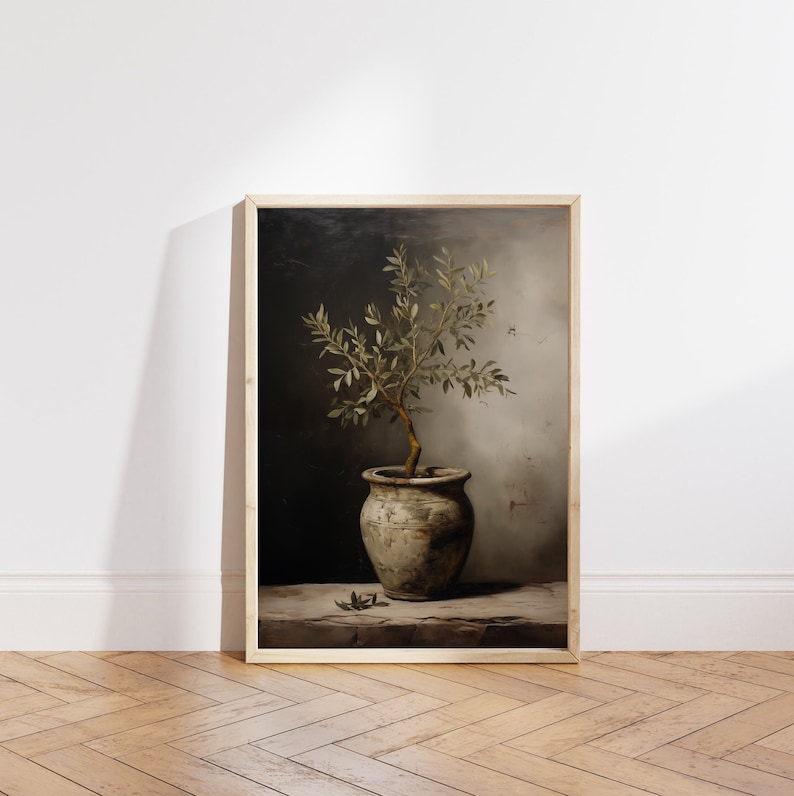 May include: A framed art print showing a potted olive tree. The image features a small tree with green leaves in a weathered, grey ceramic pot. The background is a dark, textured wall with a wooden frame.