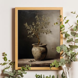 Op de afbeelding: Een ingelijste kunstdruk met een olijfboom in pot. Het kunstwerk toont een kleine boom met groene bladeren en een bruine stam in een verweerde, grijze keramische pot. De achtergrond heeft een donkere, sombere esthetiek.