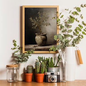 May include: A framed botanical print of a potted plant, surrounded by decorative elements. Eucalyptus branches in a white jug, small cacti in terracotta pots, and a vintage camera are arranged on a wooden surface.
