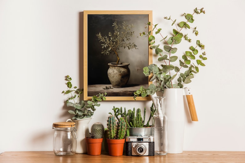 May include: A framed botanical print of a potted plant, surrounded by decorative elements. Eucalyptus branches in a white jug, small cacti in terracotta pots, and a vintage camera are arranged on a wooden surface.
