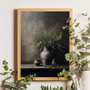 May include: A framed print of a still life featuring a gray ceramic vase with a bouquet of green foliage, two smaller gray ceramic pots, and a wooden table. The background is a muted gray with a soft, blurred effect.