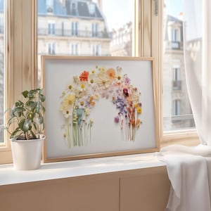 Puede incluir: Una obra de arte floral enmarcada con flores prensadas que forman un arcoíris. Las flores están dispuestas en degradado, desde el amarillo hasta el morado. La obra se exhibe en un alféizar de ventana con una planta en maceta en primer plano.