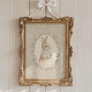 May include: A framed print featuring a watercolor illustration of a bunny wearing a pink dress, set within a floral wreath. The ornate gold frame is suspended by a white ribbon bow against a white background.