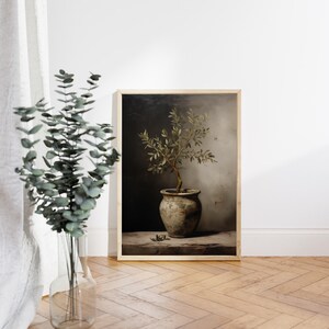 May include: A framed print of an olive tree in a pot, with a vintage feel. The artwork uses a muted colour scheme. The olive tree is in a rustic pot, set against a dark background. The frame is a light wood colour.