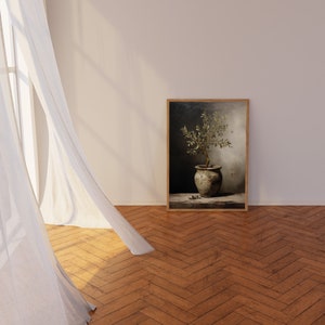 May include: A framed print depicting an olive tree in a weathered pot. The artwork is set against a dark backdrop, within a wooden frame. The image rests on a wooden floor, with white curtains in the foreground.