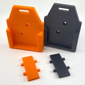 Puede incluir: Dos soportes de pared impresos en 3D, uno naranja y otro negro, con una peque&ntilde;a pieza rectangular que encaja en el soporte. Las piezas est&aacute;n dise&ntilde;adas para sujetar un dispositivo.