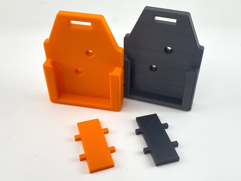 Puede incluir: Dos soportes de pared impresos en 3D, uno naranja y otro negro, con una peque&ntilde;a pieza rectangular que encaja en el soporte. Las piezas est&aacute;n dise&ntilde;adas para sujetar un dispositivo.