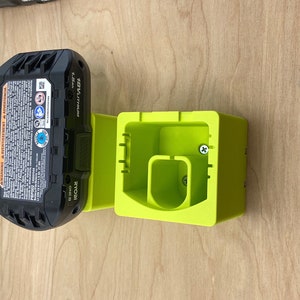 May include: A black and green 18V Lithium Ryobi battery pack attached to a bright green battery holder. The battery pack has a label with safety warnings and technical specifications. The holder is designed to receive the battery.