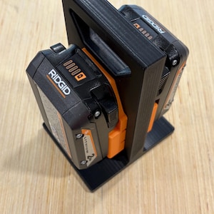May include: Black 3D-printed battery holder with two black and orange Ridgid lithium-ion batteries. The holder has a handle and a base for stability. The batteries are labeled with the Ridgid brand.
