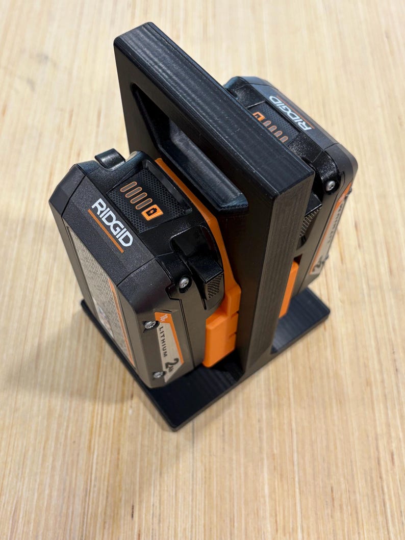 May include: Black 3D-printed battery holder with two black and orange Ridgid lithium-ion batteries. The holder has a handle and a base for stability. The batteries are labeled with the Ridgid brand.