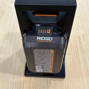 May include: A black 3D-printed battery holder with a Ridgid power tool battery. The battery is black and orange, and the holder has a rectangular design with a handle-like opening at the top.