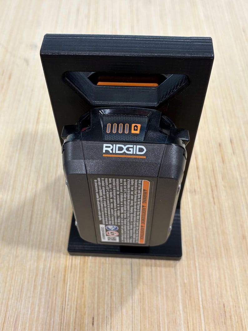 May include: A black 3D-printed battery holder with a Ridgid power tool battery. The battery is black and orange, and the holder has a rectangular design with a handle-like opening at the top.