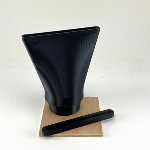 Puede incluir: Un embudo negro impreso en 3D con una abertura rectangular y una base redonda, sentado sobre una base de madera con una varilla negra impresa en 3D.