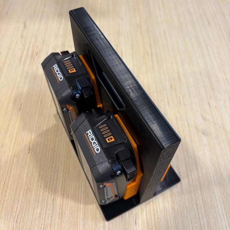 May include: Black 3D-printed power tool battery holder designed to hold two Ridgid brand batteries. The holder features a rectangular base and a vertical back with slots for the batteries. The batteries are black and orange.