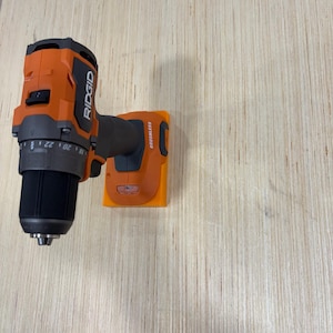 May include: An orange and black Ridgid cordless drill with a black drill bit. The drill is mounted on an orange holder. The drill has the word "RIDGID" printed on the side.