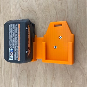 Puede incluir: Un soporte de pared para cargador de bater&iacute;a negro y naranja con dos tornillos. El cargador de bater&iacute;a est&aacute; etiquetado como "2Ah Li-Ion Battery".